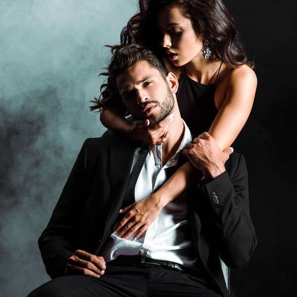 passionate woman standing and hugging handsome bearded man on black with smoke 