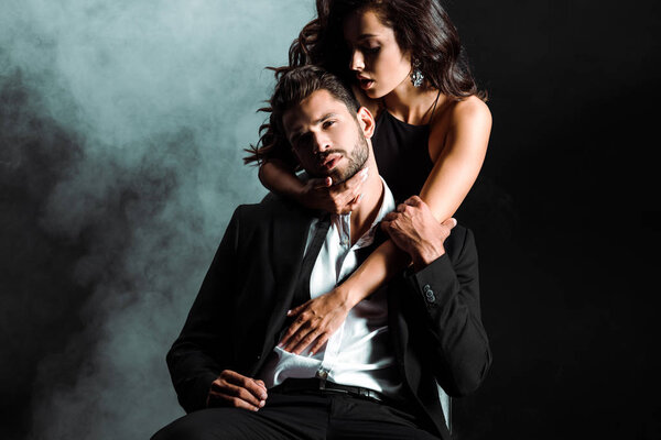 passionate young woman standing and hugging handsome bearded man on black with smoke 
