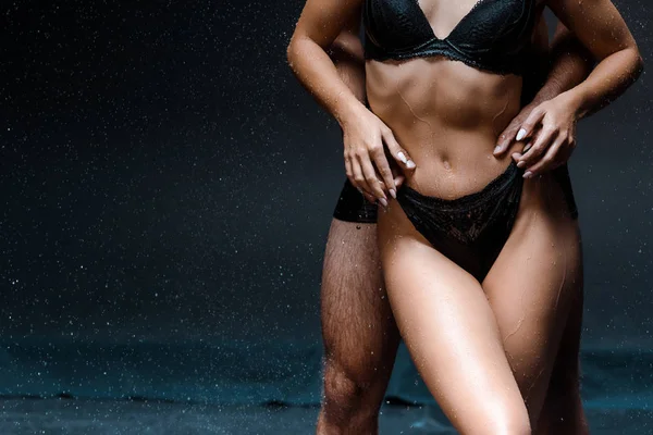 cropped view of muscular man hugging young wet woman under raindrops on black