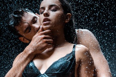 low angle view of shirtless and wet man touching face of attractive girlfriend with closed eyes under raindrops on black  clipart