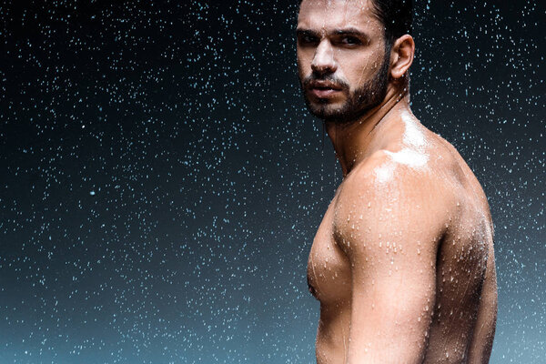 wet man looking at camera while standing under raindrops on black 