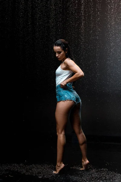 sexy young and wet woman standing on tip toe under raindrops on black