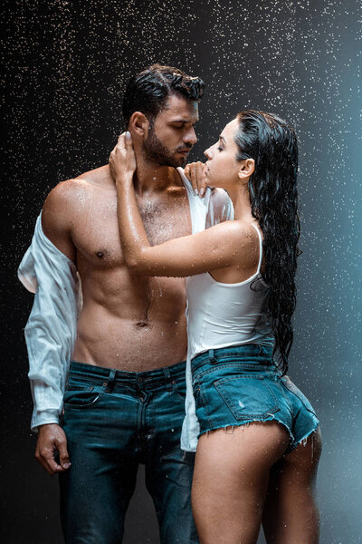 undressed and muscular man standing with sexy girlfriend under raindrops on black 