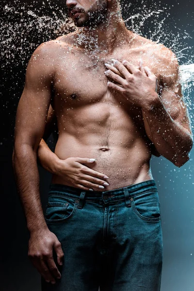 cropped view of woman hugging wet bearded man in denim jeans near water splash on black