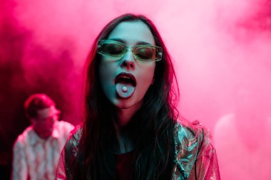 beautiful girl in sunglasses with lsd on tongue in nightclub with pink smoke clipart
