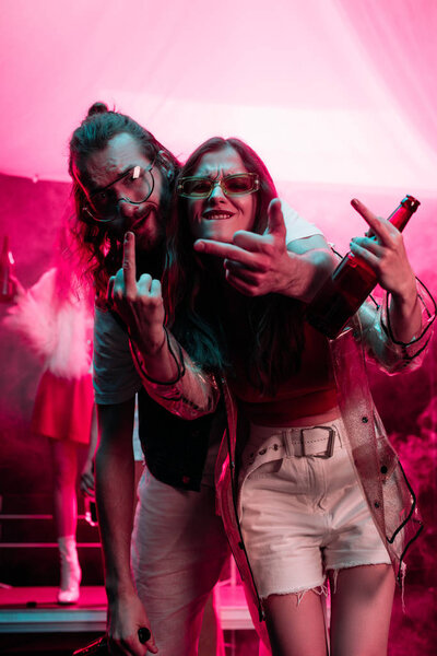 young woman with beer and handsome man showing middle fingers in nightclub during rave 