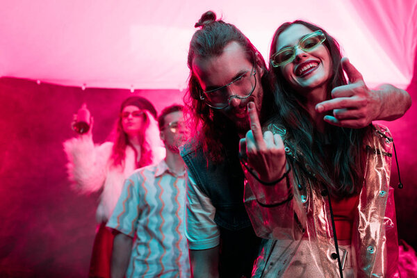 happy girl and man in sunglasses showing middle fingers in nightclub during rave party 