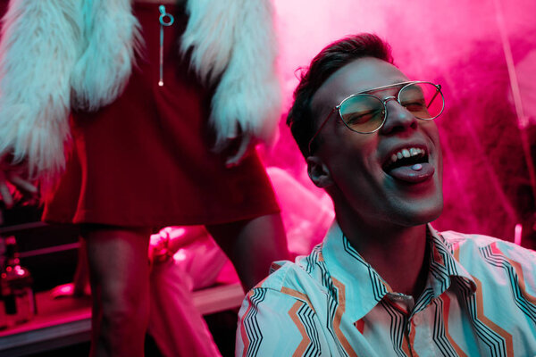 девушка рядом с мужчиной в солнцезащитных очках с lsd на языке в ночном клубе с розовым дымом
