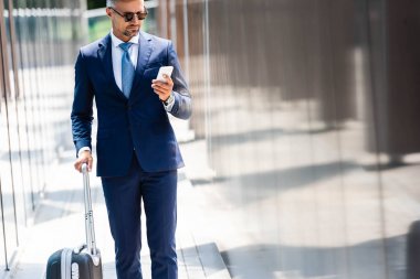 takım elbise ve gözlük yakışıklı işadamı akıllı telefon kullanarak ve bavul tutan