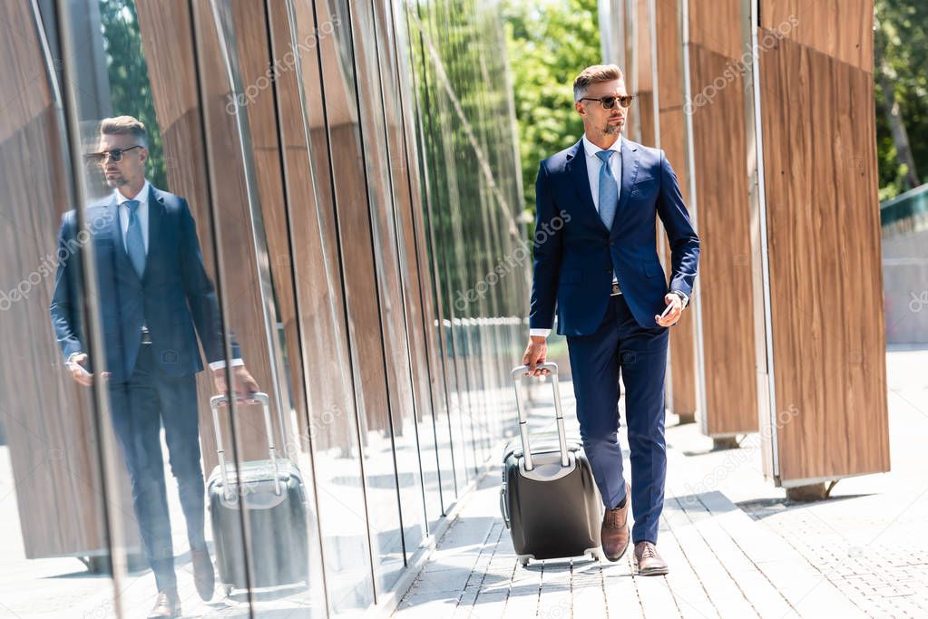 handsome businessman in suit and glasses holding smartphone and suitcase