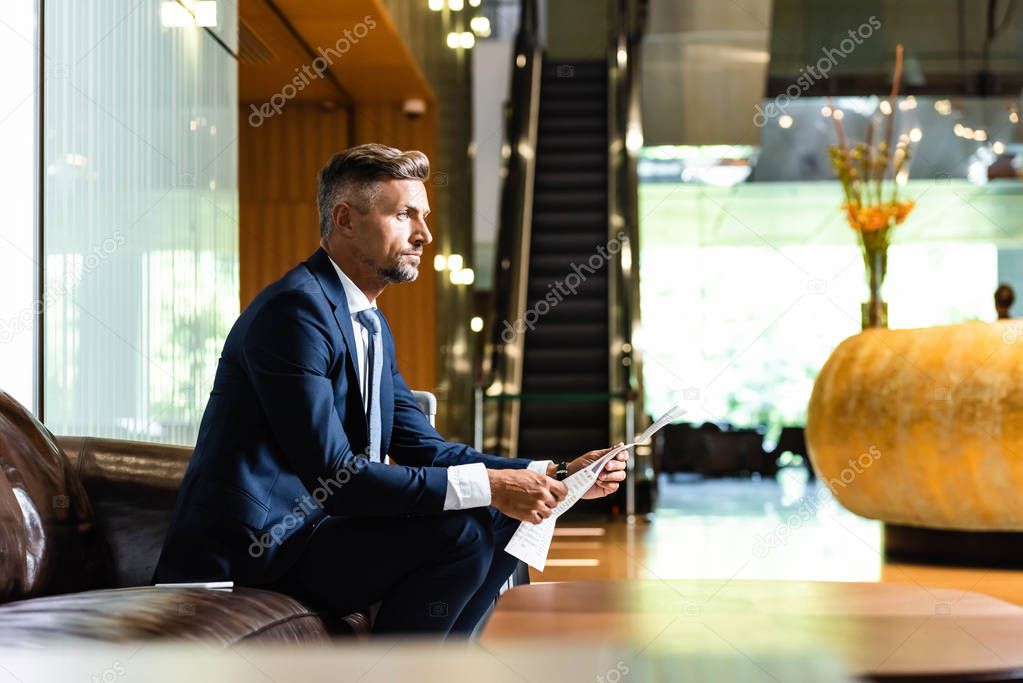 side view of handsome businessman in suit sitting on sofa and holding newspaper 