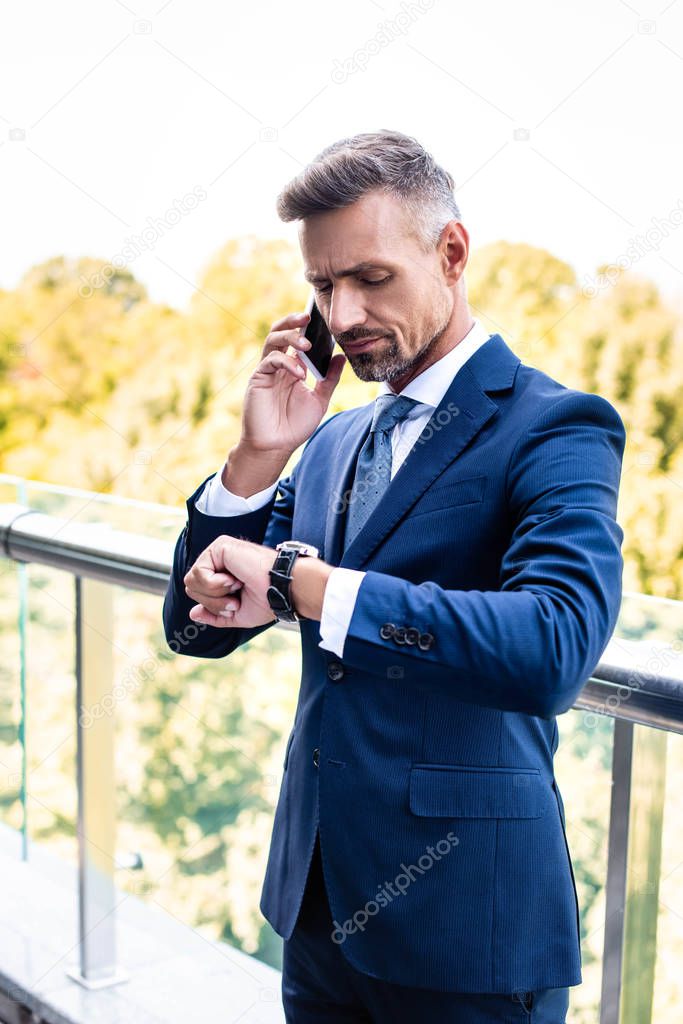 handsome businessman in formal wear talking on smartphone and looking at watch 