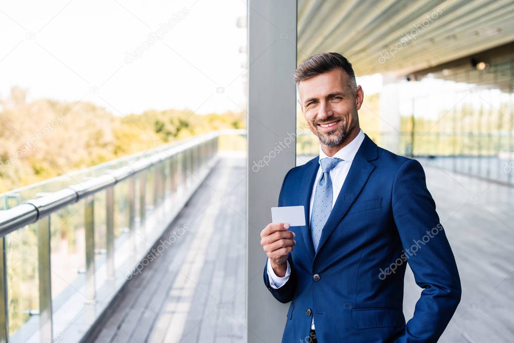 handsome businessman in formal wear with hand in pocket holding blank card 
