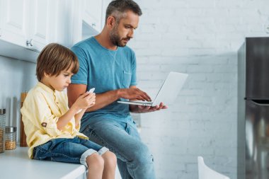 smarthone kullanarak oğlu yakınında mutfak tezgahında otururken dizüstü bilgisayar kullanan adam 