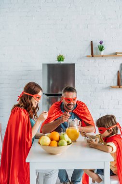 family in costumes of superheroes having breakfast in kitchen together 
