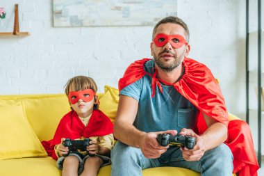 Kiev, Ukrayna - 5 Temmuz 2019: Evde kanepede otururken video oyunu oynayan süper kahraman ların kostümlü baba ve oğlu