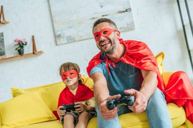 Kiev, Ukrayna - 5 Temmuz 2019: Heyecanlı baba ve oğul süper kahraman kostümleri kanepede oturan ve video oyunu oynarken