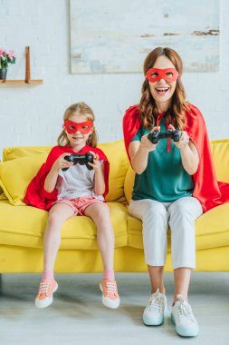 Kiev, Ukrayna - 5 Temmuz 2019: Evde kanepede otururken video oyunu oynayan süper kahraman kostümleri mutlu anne ve kızı
