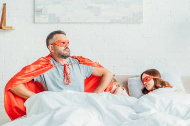 serious father in costume of superhero sitting in bed near sleeping family, holding hands on hips and looking away clipart