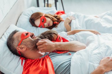 selective focus of man in superhero costume scratching cheek while lying in bed near wife and son clipart