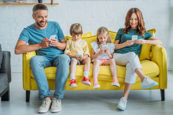 happy mother and father with children sitting on sofa and using smartphones
