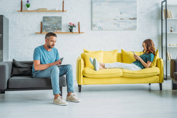 woman lying on yellow sofa with smartphone near husband sitting on couch and using smartphone