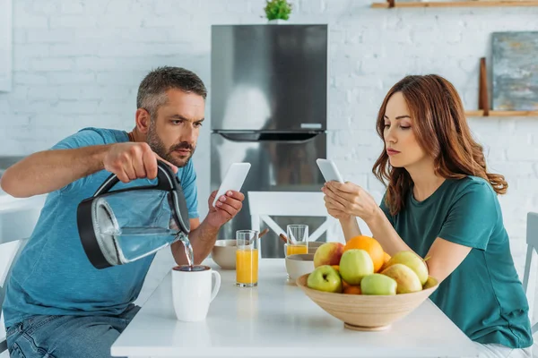 スマートフォンを使用して妻の近くの台所に座っている間 カップに水を注ぐスマートフォンを持つ男 — ストック写真