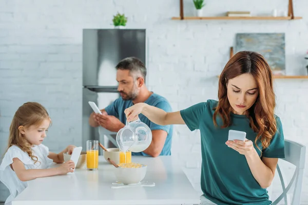 妇女与智能手机过度填充碗牛奶 而坐在厨房的桌子附近丈夫和女儿使用智能手机 — 图库照片