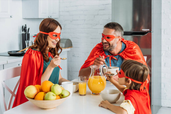 cheerful family in costumes of superheroes having breakfast in kitchen