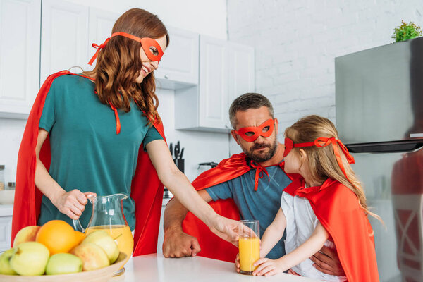 smiling mother, father and daughter in costumes of superheroes drinking orange juice 