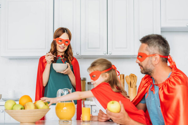 family in costumes of superheroes having breakfast in kitchen together