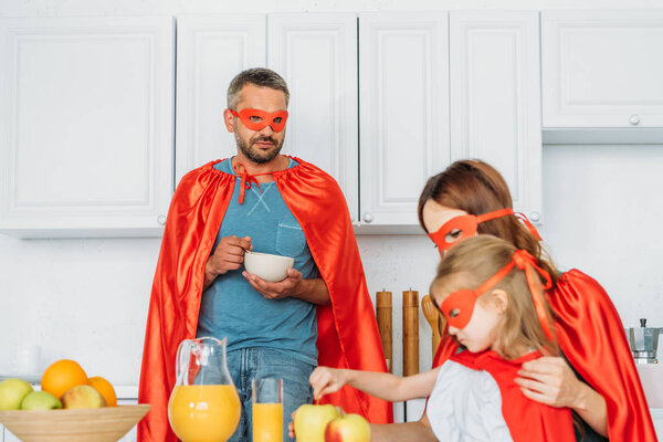 father, mother and daughter in costumes of superheroes having breakfast in kitchen 