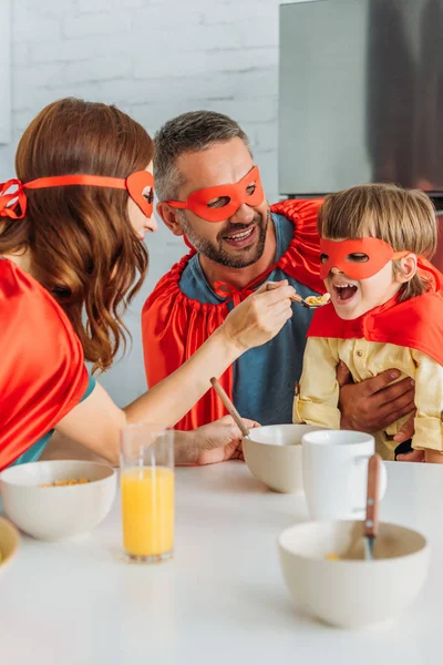 欢快的家庭在超级英雄的服装有早餐 而母亲喂养与薄片的儿子 — 图库照片