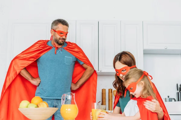 父親が腰に手を置きながら台所で朝食を食べているスーパーヒーローの衣装を着た家族 — ストック写真