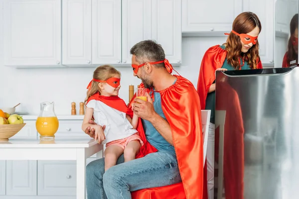 身着超级英雄服装的家庭在厨房里度过时光 而父亲和女儿喝橙汁 母亲站在冰箱附近 — 图库照片