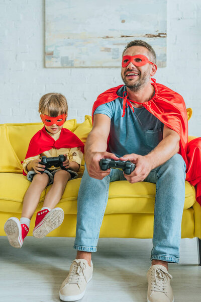 KYIV, UKRAINE - JULY 5, 2019: Cheerful father and son in costumes of superheroes sitting on sofa and playing video game