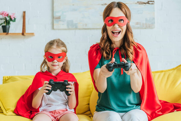 KYIV, UKRAINE - JULY 5, 2019: Cheerful mother and daughter in costumes of superheroes playing video game while sitting on sofa at home