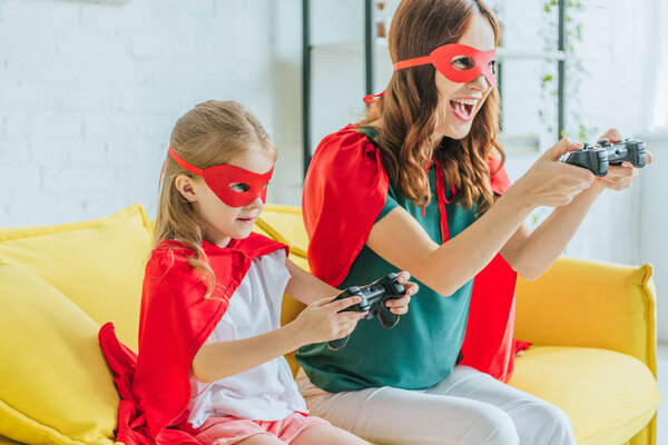 KYIV, UKRAINE - JULY 5, 2019: Excited woman and cute chid in costumes of superheroes playing video game at home
