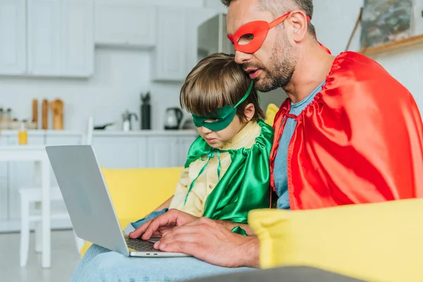 selective focus of dad and son in superheroes costumes using laptop at home