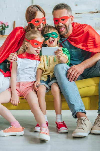 欢快的家庭在超级英雄服装坚持舌头出来 而采取自拍在智能手机在家里 — 图库照片