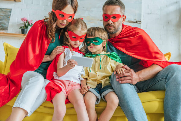 parents with two kids in costumes of superheroes using smartphone together