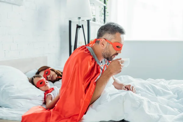 father in costume of superhero drinking coffee while sitting in bed near sleeping wife and son