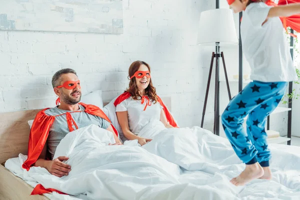 Cheerful Parents Superheroes Costumes Looking Son Jumping Bed — Stock Photo, Image