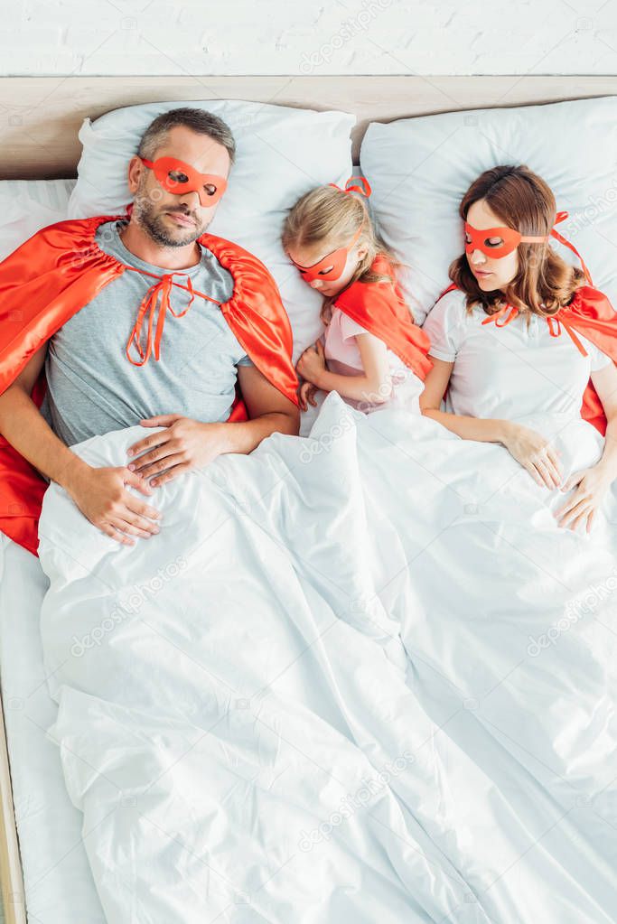 top view of family sleeping on white bedding in costumes of superheroes 