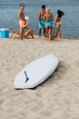 white surfing board on sand near multicultural friends resting on beach clipart