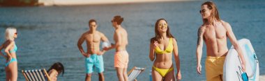 panoramic shot of young man holding surfing board near smiling girl in swimsuit while multicultural friends resting on beach clipart