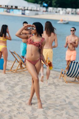 beautiful african american woman in swimsuit looking away while standing on beach near multicultural friends clipart
