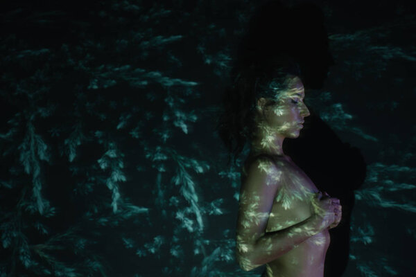 attractive girl with closed eyes covering breasts near pines on black 