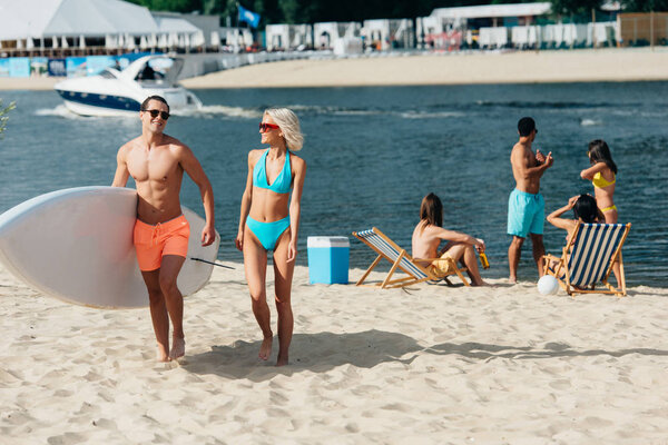 smiling man holding surfboard while walking near attractive woman on beach