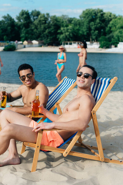 smiling multicultural friends looking at camera while sitting on beach in chaise lounges and holding bottles with beer